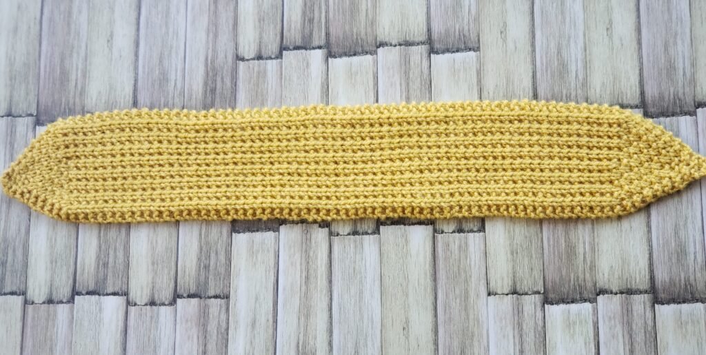 Full-length horizontal view of an un-tied mustard yellow knit headband showing symmetrical tapered points and garter stitch borders