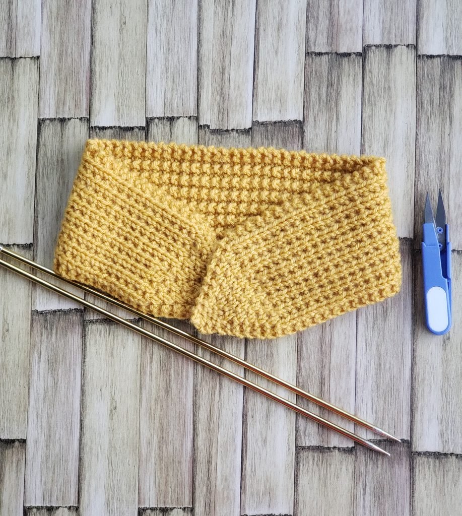 Mustard yellow knitted tie headband laid flat on a rustic wood surface with brass knitting needles and blue yarn snips