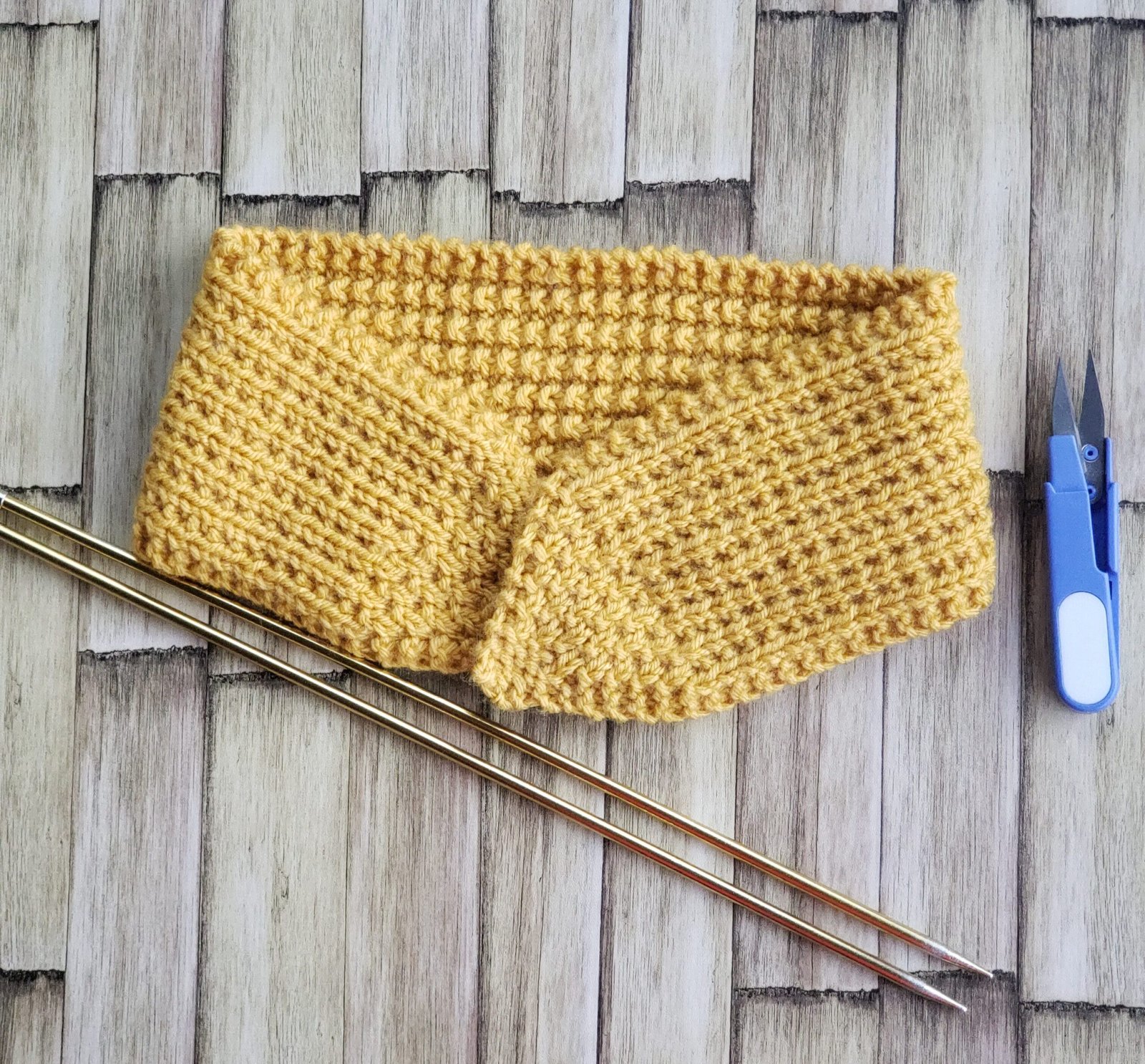 Mustard yellow knitted tie headband laid flat on a rustic wood surface with brass knitting needles and blue yarn snips